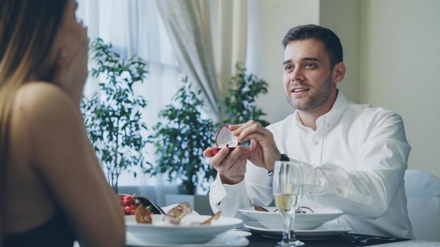 Happy loving man in white shirt is proposing to surprised beautiful girlfriend, then giving her engagement ring during romantic date in restaurant. Romance and relationship concept.
