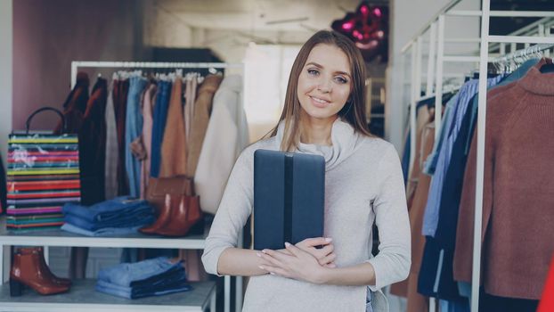 Portrait of young businesswoman standing in her clothing boutique, holding tablet, smiling and looking at camera. Spacious store with women's clothes in background. Successful start-up concept.