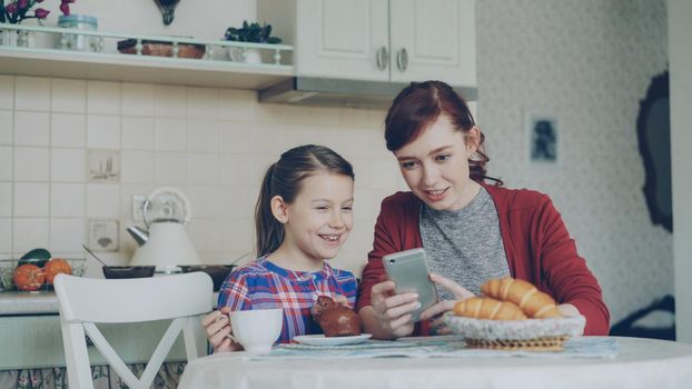 Cheerful mother and little daughter browsing smartphone together and smiling during breakfast in the morning in kitchen at home