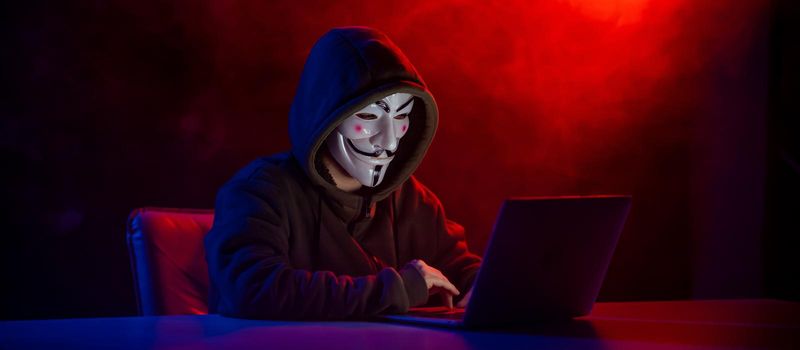 June 5, 2022 Novosibirsk, Russia: Anonymous in a hood is typing on a laptop in the dark in red-blue smoke