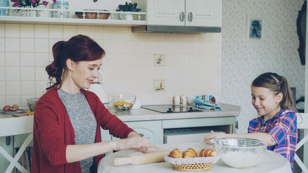 Young mother rolling dough and talking to her little cute daughter while cooking in the kitchen on weekend. Family, food, home and people concept