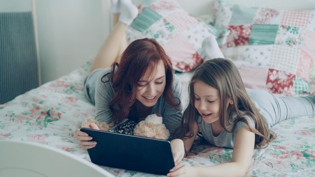 Attractive young mother and her cute daughter in pajamas laughing and looking in digital tablet while lying on bed at home in the morning