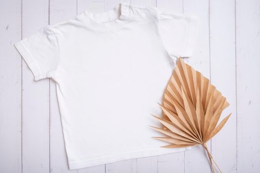 White children's t-shirt mockup for logo, text or design on wooden background with palm leaves top view.