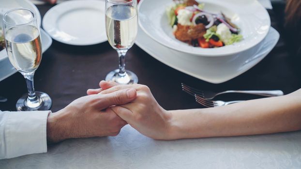 Close-up shot of young lovers touching and holding hands at romantic dinner in classy restaurant. Table with sparking champagne glasses, flatware and food in background.