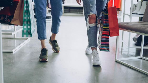women's legs walking slowly through luxurious shop. Women are wearing jeans and trainers and carrying bright paper bags. Shopaholic lifestyle concept.