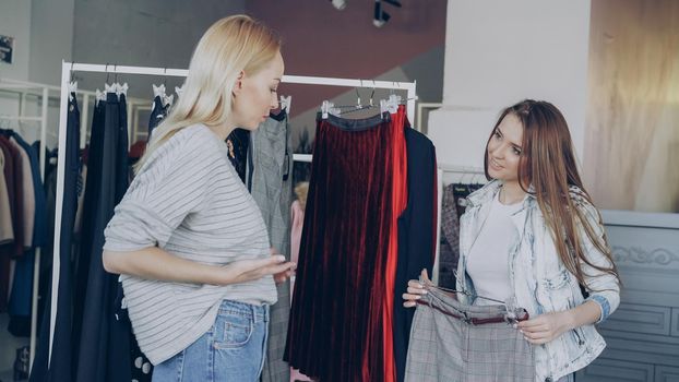 Cheerful pretty girls are choosing trousers together while shanding in nice clothes boutique. They are taking stylish pants, checking fabric, length and belt, fitting them and discussing purchase