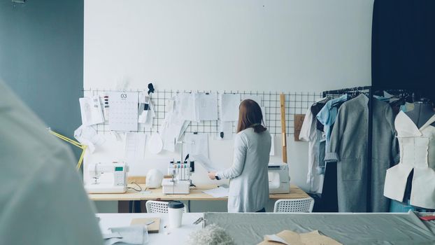 Back view of creative female tailor taking sketches from tailoring desk and hanging them on white wall above table. Planning, designing and creating new collection concept.