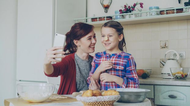 Smiling mother together with funny cute daughter taking selfie photo with smartphone camera making silly face while cooking at home in kitchen. Family, cook, and people concept