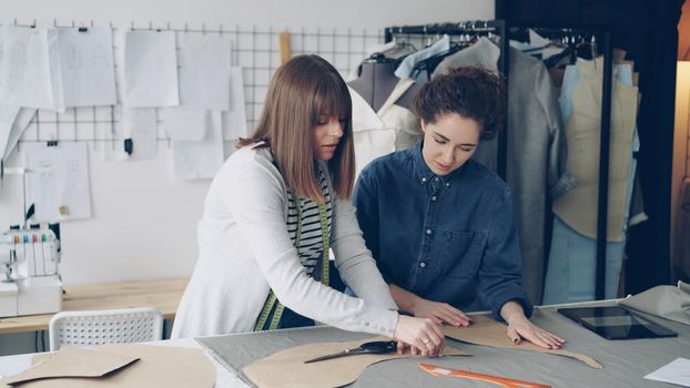 Team of creative designers is working with clothing patterns and fabric in tailor's shop. Young women are concentrated on measuring. Multiple sewing items and tools in background.