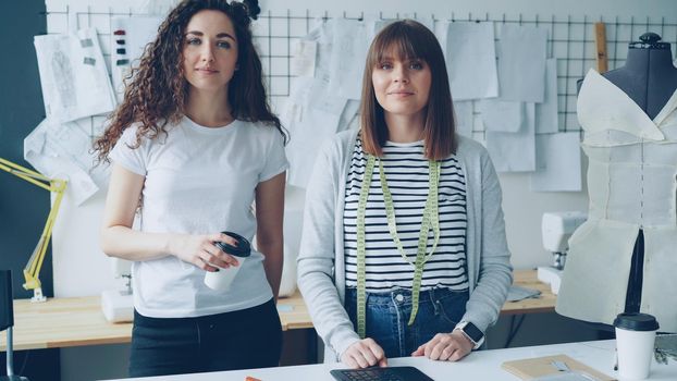 Portrait of two young female clothing designers entrepreneurs standing in workshop together and looking at camera. One woman is holding take-out coffee, other is touching studio desk.