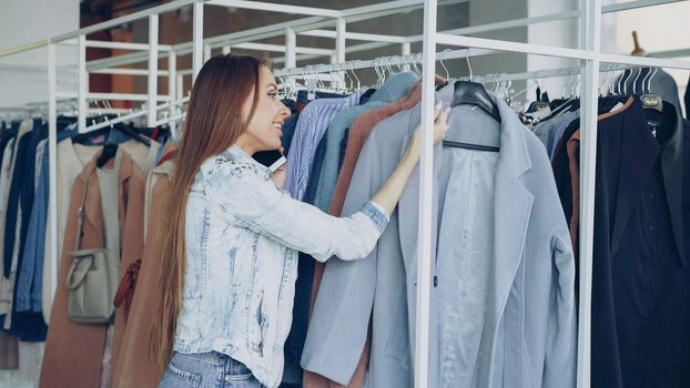 Pretty young woman is talking on mobile phone and shopping for clothes in luxurious boutique. She is looking at fashionable coat carefully, touching it and smiling with pleasure