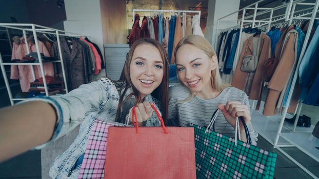 Point of view shot of two attractive careless girls making selfie with colourful paper bags in women's clothes shop. Friends are posing together, laughing and chatting happily