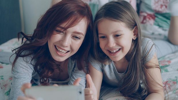 Happy mother and little girl taking selfie photo with smartphone camera and have fun grimacing while sitting in cozy bedroom at home