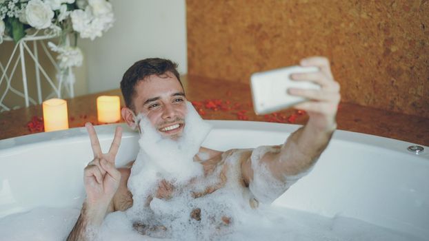 Handsome cheerful guy is taking selfie in bathtub with soap foam on his beard using smart phone. He is laughing and making gestures with his hand posing and having fun.