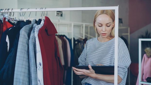 Young businesswoman is checking garments on rails and working with tablet in her clothing shop. She is checking prices and labels, touching screen and typing. Successful start-up concept.