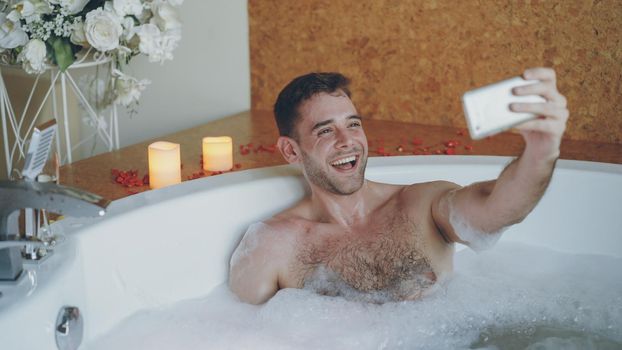 Young handsome man popular blogger is recording video in hot tub in day spa using smartphone for his blog. Burning candles, champagne glass and flowers are visible.