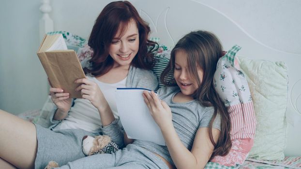 Careful mother helping her little cute daughter with homework for elementary school. Loving mom reading a book and girl writing notes in copybook while sitting together in cozy bedroom at home
