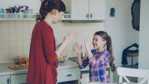 Happy young mother and cute funny daughter playing clapping hands game and have fun while cooking in kitchen at home. Family, cooking and people concept