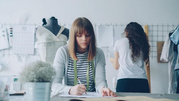 Young attractive clothing designer is busy drawing sketch with pencil while her colleague is watching pictures pinned on wall in design studio. Creative thinking concept.