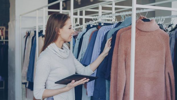 Young and successful owner of women's clothing shop is checking and counting garments on rails while using tablet. She is typing information about her goods. Small business concept.