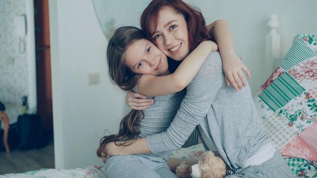 Portrait of cute smiling girl embracing her happy mother and looking at camera together while sitting on bed in bright bedroom at home