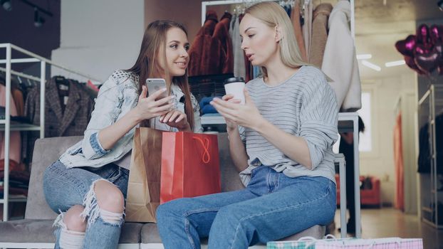 Attractive girls are sitting together in women's clothing shop. They are drinking coffee, chatting carelessly and using smartphone. Nice boutique, colourful garments and baloons in background.