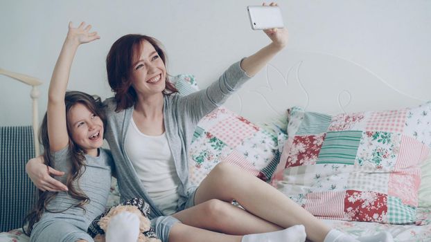 Happy mother and little girl taking selfie photo with smartphone camera and have fun grimacing while sitting in cozy bedroom at home