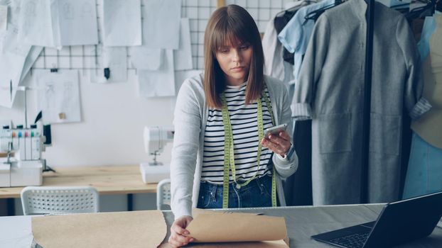 Clothing design entrepreneur is checking paper sewing drafts and looking at smartphone. She is examining every cutout and thinking about future garment. Modern technology in sewing industry concept.