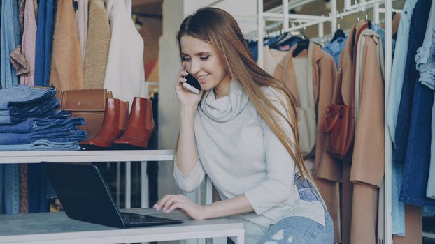 Young businesswoman working with laptop and talking cheerfully on her mobile phone sitting in clothes shop. Stylish clothing on display and customer moving in background. Small business concept.