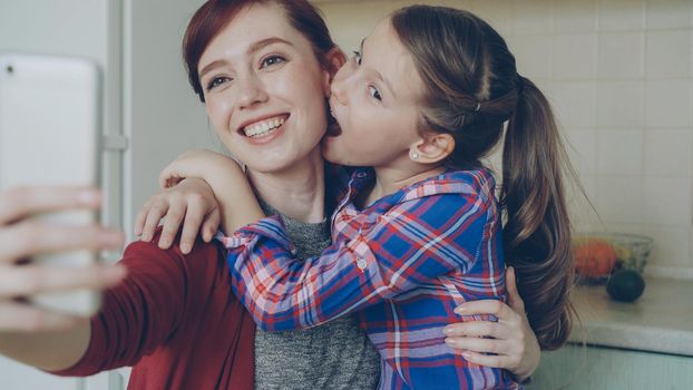 Smiling mother together with funny playful daughter making selfie photo with smartphone camera at home in kitchen. Family, cooking, and people concept