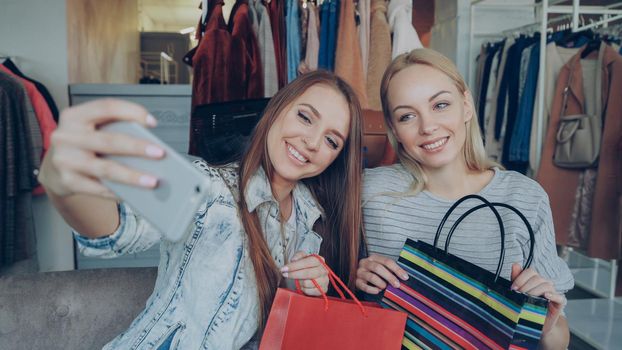 Close-up shot of two young women making selfie with smart phone after shopping in women's clothing store. First they are posing with paper bags and laughing, then watching photos on screen