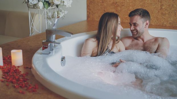 Young attractive people are hugging and talking in hot bubbling bathtub with foam in modern day spa. Romantic relationship, conversation and wellness concept.