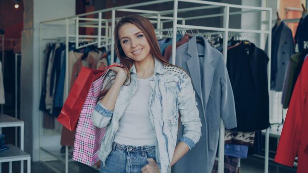 Portrait of happy and attractive girl standing in clothing shop with colourful bags, smiling gladly and looking at camera. Modern clothes is hanging in background.