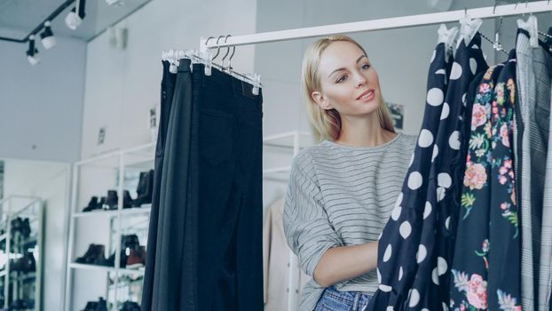 Pretty blond girl is choosing fashionable clothes in boutique. She is looking through fine garments and taking polka-dotted dress from the rails, examining its quality and length.