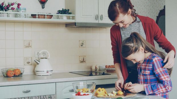 Young pretty mother teaching her cute daughter to cut vegetables properly. Little smiling girl cooking together with loving mom at home in modern kithcen