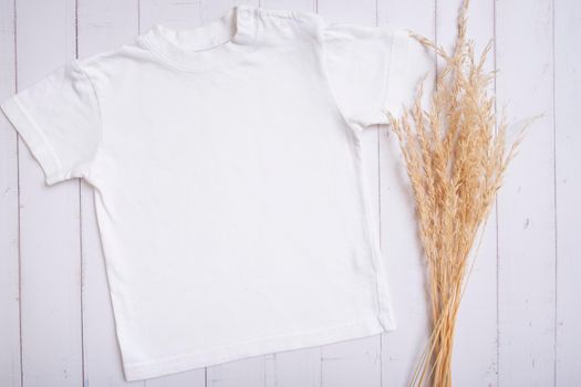 White children's t-shirt mockup for logo, text or design on wooden background with pampas grass top view.