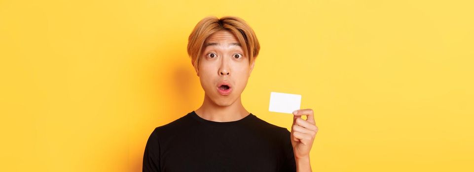 Close-up of surprised gasping asian guy with fair hair, looking amazed and startled as showing credit card, standing yellow background.