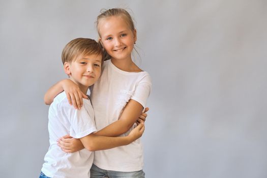 Cute two kids, little boy and girl in white t-shirts hugging, smiling and looking at the camera on gray background