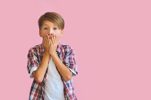 Young surprised boy dressed in jeans, a white T-shirt and a plaid shirt covers mouth with palms, isolated on pink background with copy space