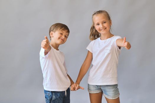 Cute two kids, little boy and girl in white t-shirts show thumbs in air recommending, hold hands best friends isolated on gray background. Blank white t-shirt for your design