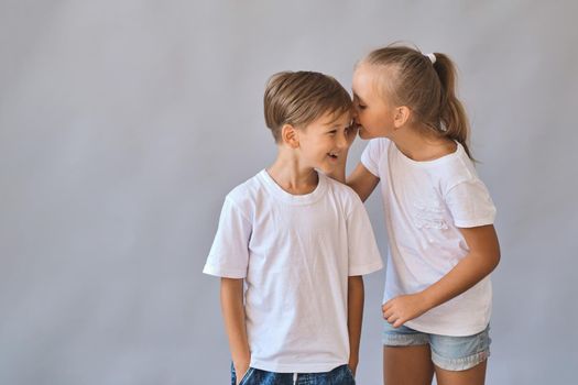 Cute two kids, little boy and girl in white t-shirts, best friends. Girl whispering secrets into boy's ear. on gray background with copy space