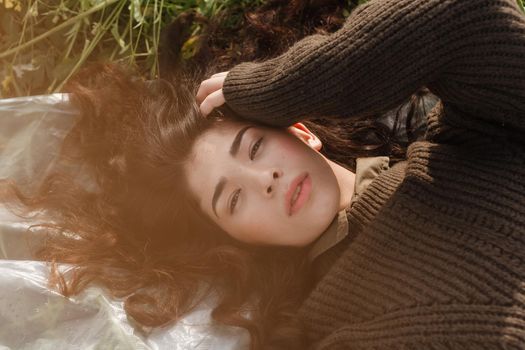 An Asian woman is lying on the ground in a field in a brown sweater. The concept of manufacturing clothing from recycled plastic. An Asian woman is lying on a plastic bag. Portrait of a dark-haired woman with loose hair.