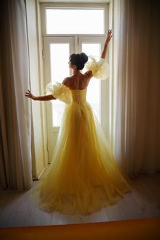 A woman's silhouette in a golden luxurious dress against the background of a window holds a curtain with her hands. Elegant lady in a yellow long silk dress with bare back, back view