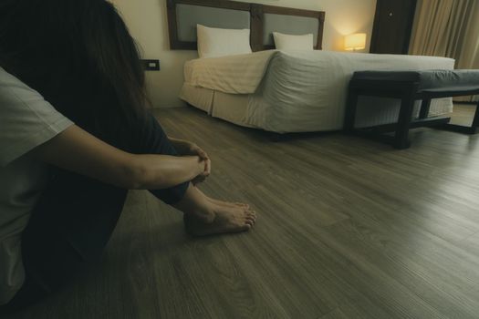 Depressed and stressed woman sitting on vinyl tiles floor near bed in hotel bedroom. Sad woman sitting in the bedroom. Girl with mental health problems. Unhappy life. World Mental Health Day concept.