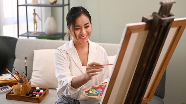 Smiling young woman painting picture on canvas with oil paints in bright cozy living room. Art, creative hobby and leisure activity concept.