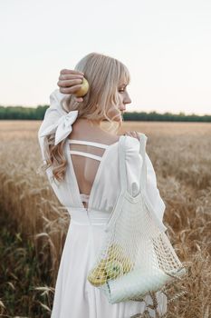 A blonde woman in a long white dress walks in a wheat field. The concept of a wedding and walking in nature.