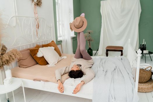 Lifestyle concept. A young middle-aged woman in a sweater lies on the bed on her back, laughs and holds a brown hat on her feet