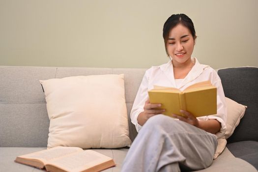 Satisfied millennial woman relaxing on couch and reading book, enjoy stress free peaceful mood in cozy living room.