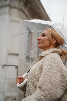 The blonde stands under a transparent umbrella during the rain. The fall season. Rear view. The woman is dressed in a black lace dress, her hair pulled back in a ponytail