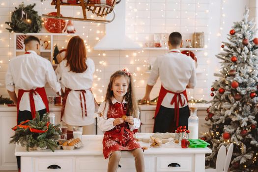 A little girl in the New Year's kitchen is sitting on the table with cookies in her hands and her parents are preparing Christmas dinner.